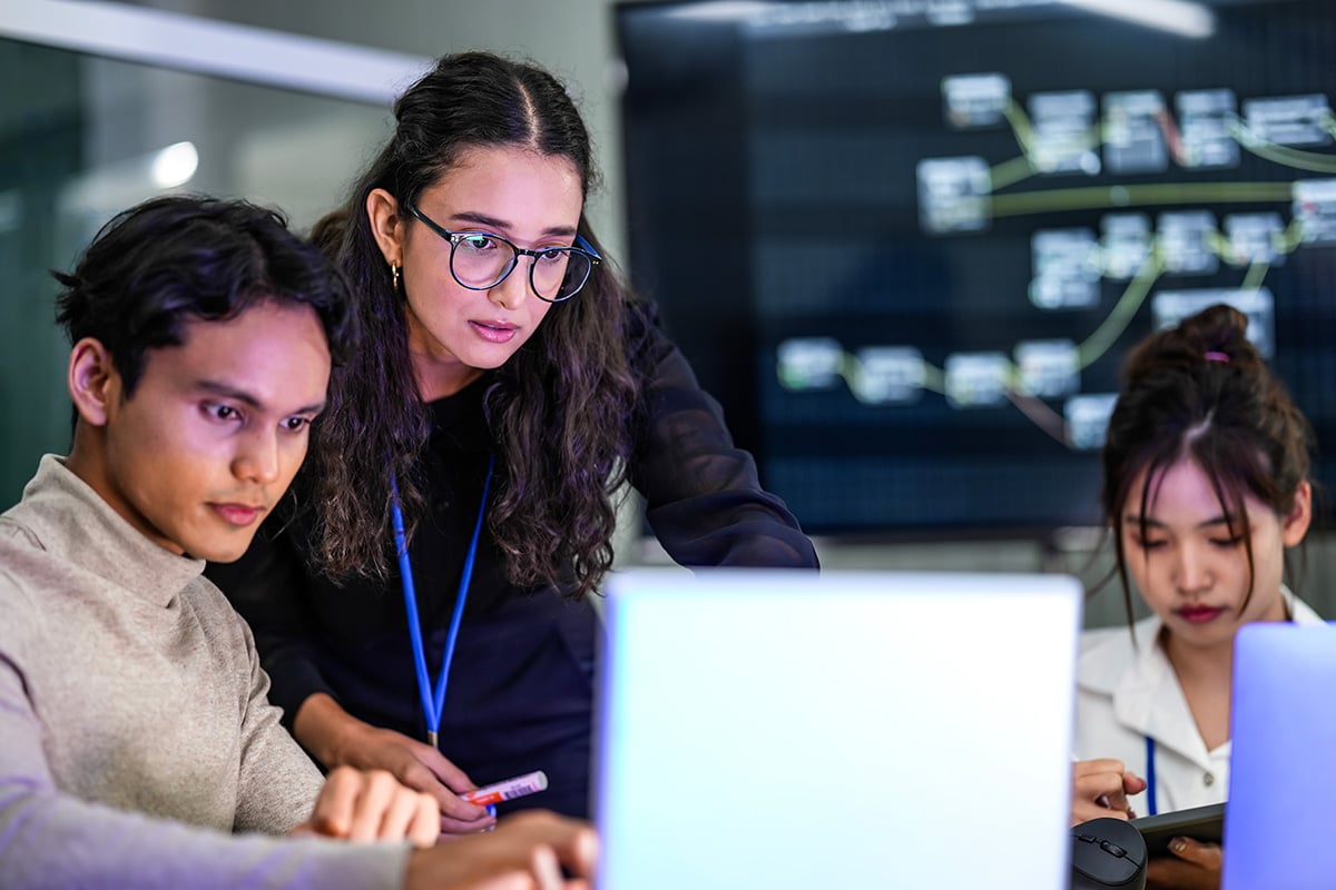 Three people collaborating around a laptop while reviewing information on a screen