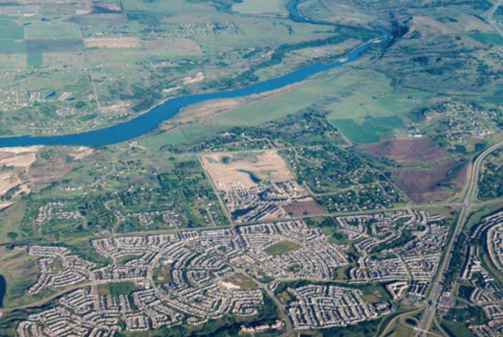Aerial view of residential neighbourhoods transitioning to farmland, with a river and highway in the background.