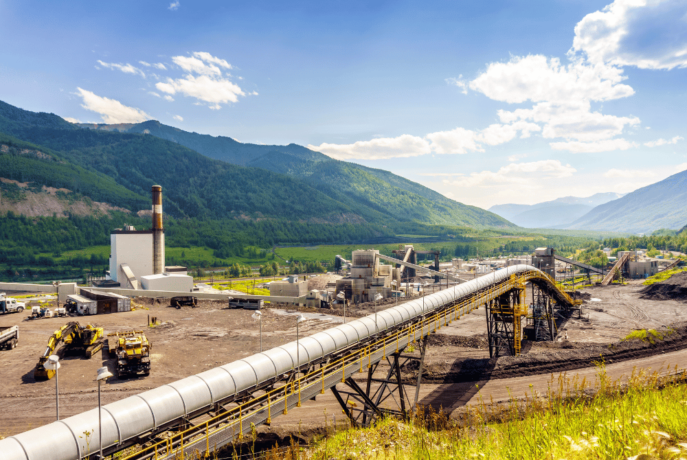 Pipeline and conveyors at a mountain valley industrial facility, with heavy equipment and a smokestack against forested peaks.