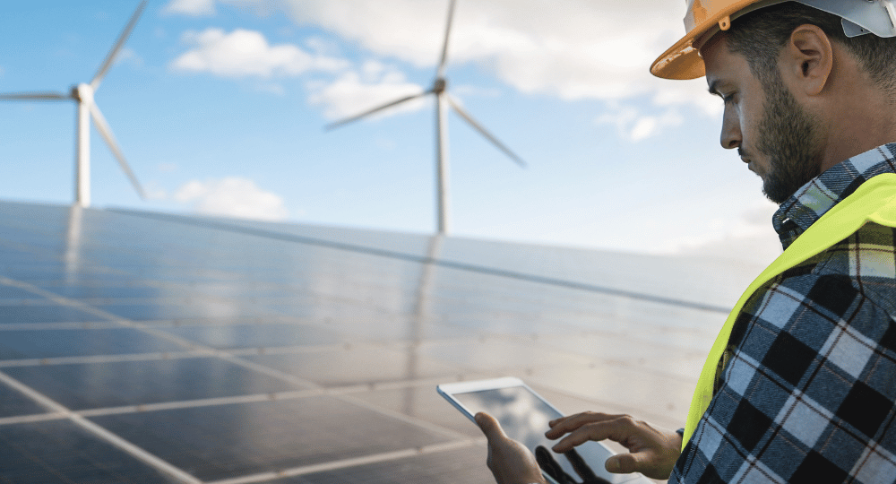 Worker using a tablet beside solar panels with wind turbines visible in the background.