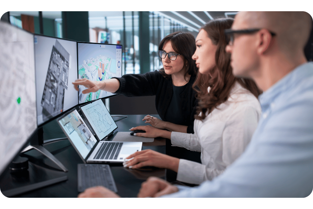 Three people collaborating at a workstation with multiple monitors displaying maps and technical graphics