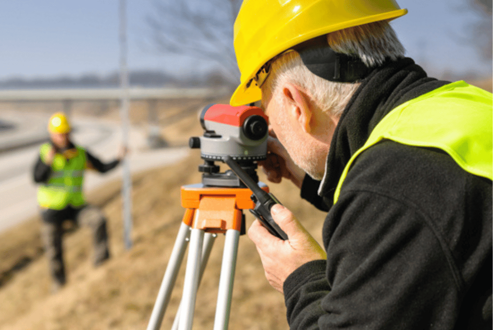 Surveyor using a leveling instrument on a tripod while another worker stands in the background