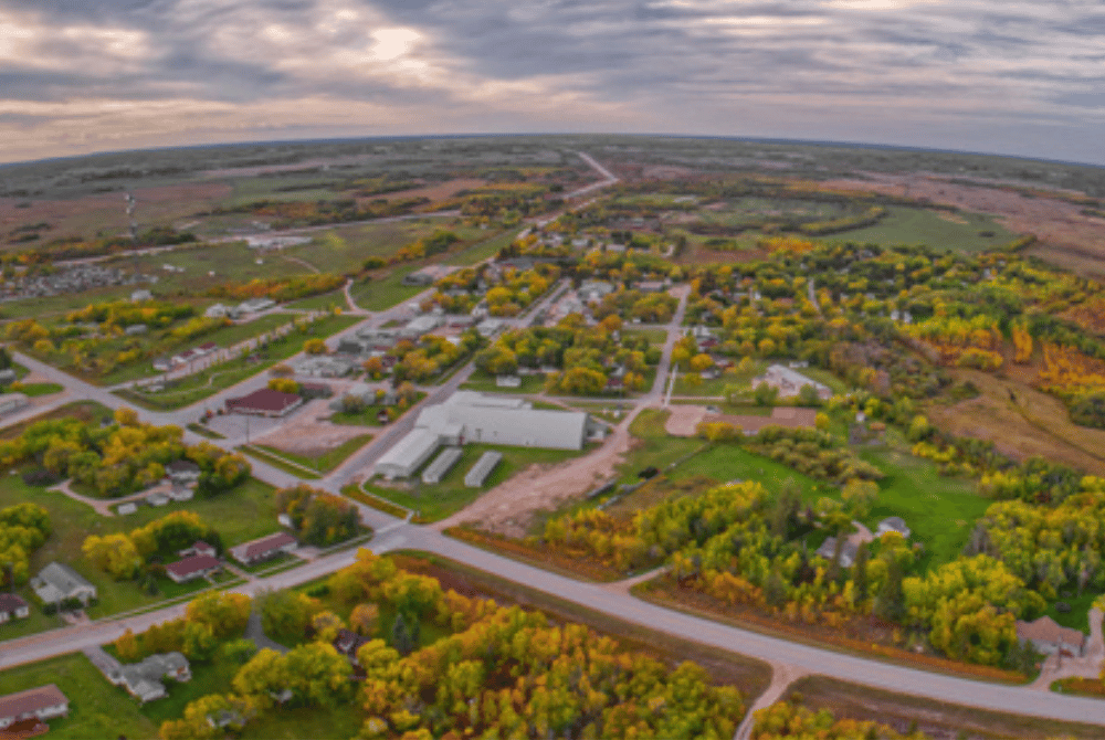 Aerial view of a small Manitoba town with residential streets, commercial buildings, and autumn foliage under an overcast sky.