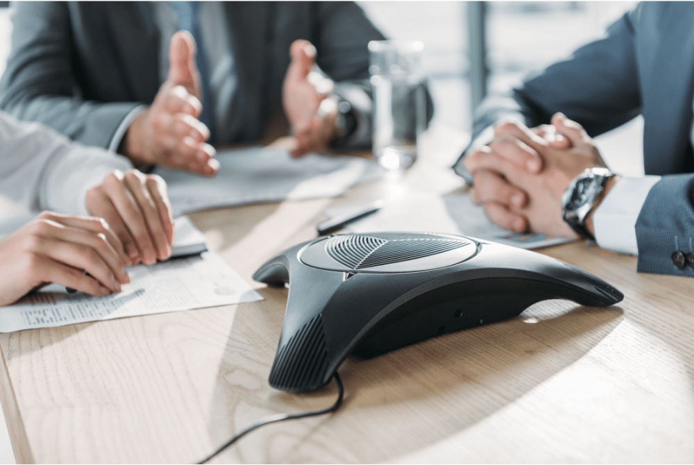 Conference phone on a meeting table surrounded by people reviewing documents and participating in a discussion