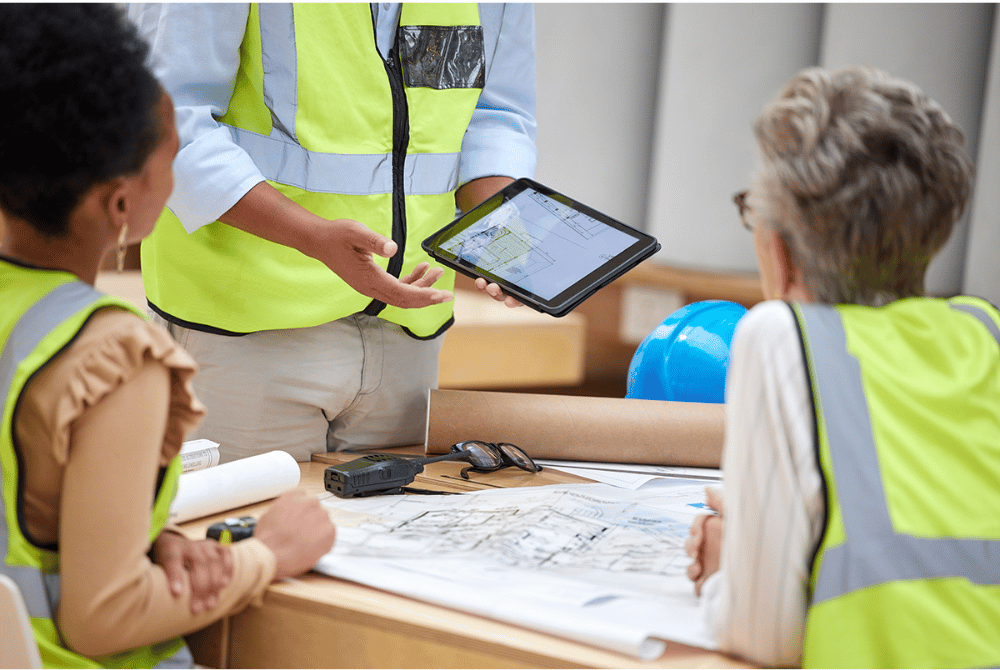 Construction team reviewing plans at a table while a standing colleague presents a digital site map on a tablet
