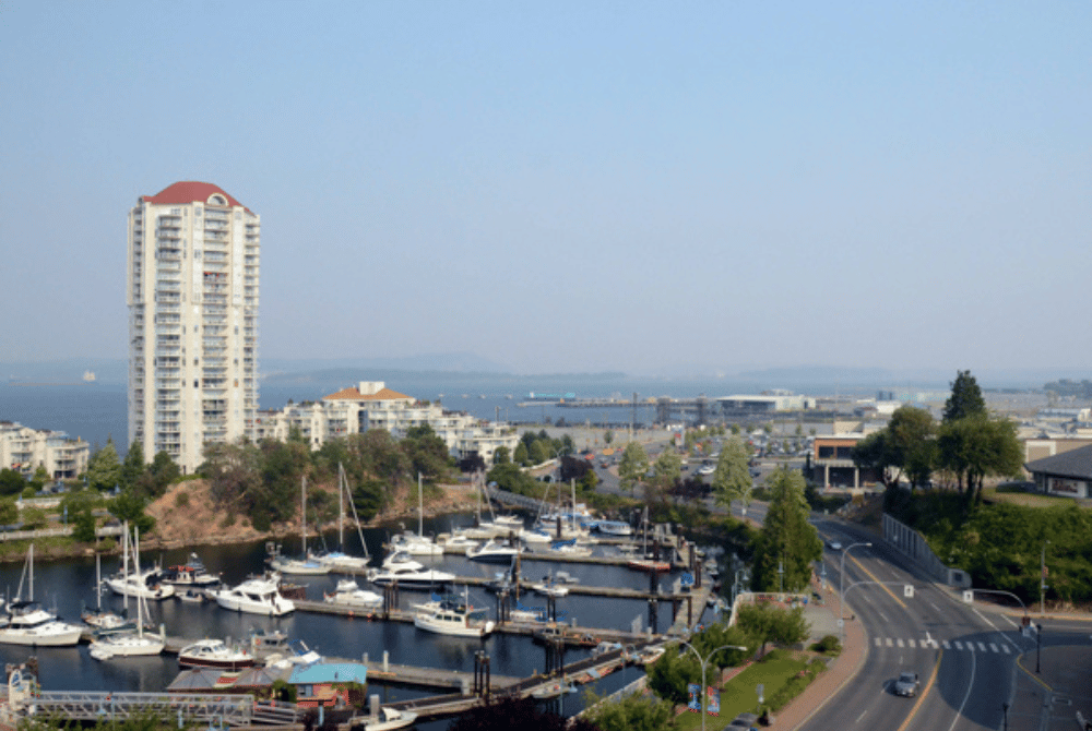 View of Nanaimo Harbour with marina docks, boats, waterfront buildings, and a residential tower overlooking the shoreline